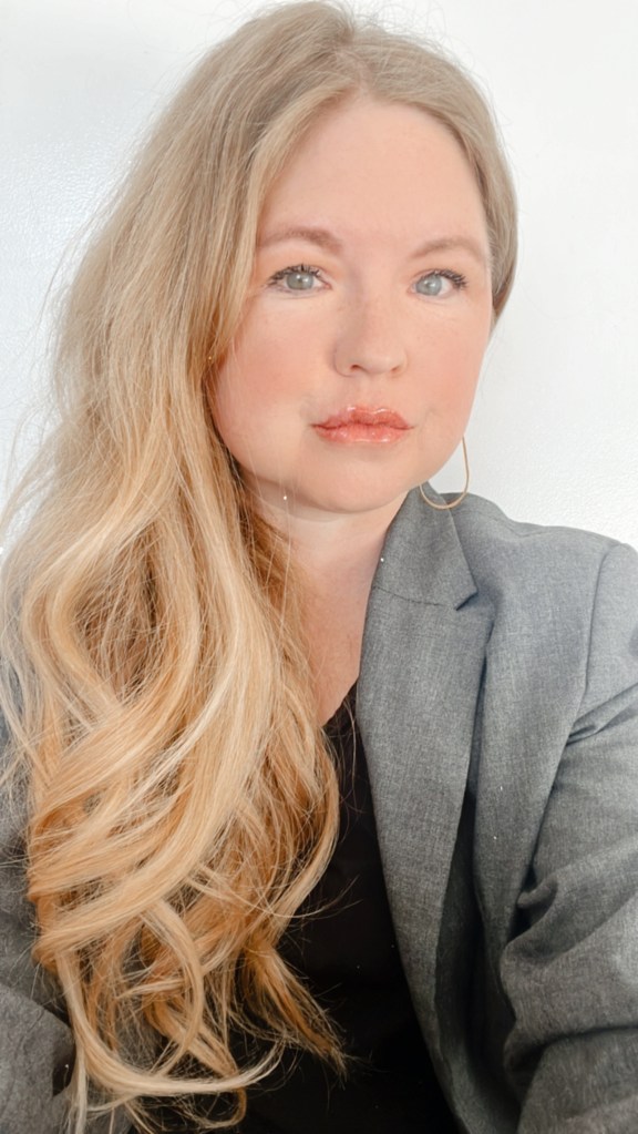 Tahnie Woodward has long blonde hair and blue-grey eyes. She is wearing a grey suit jacket and black shirt with gold hoops in her ears. She is gazing into the camera with purpose.