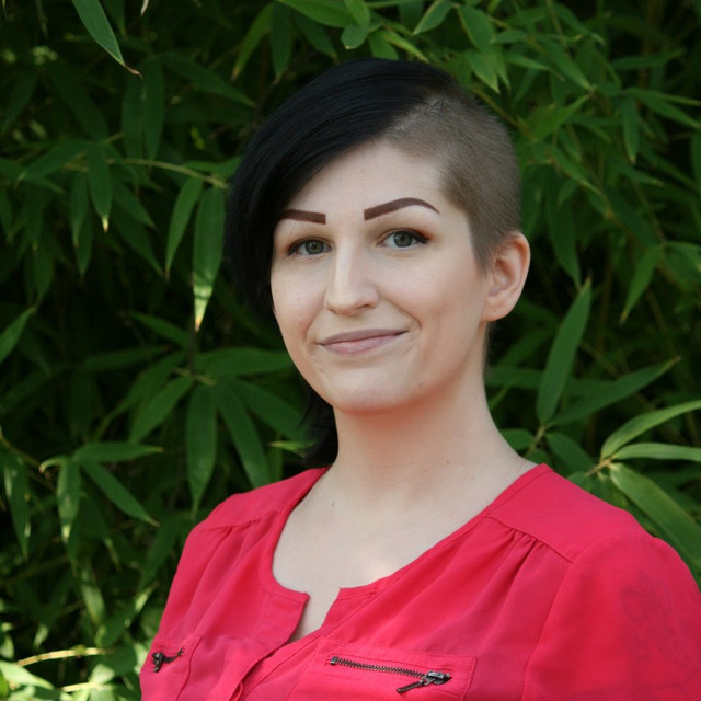 Aston Martinez uses they/them pronouns. In this image, Aston has grey green eyes and black hair with one side shaved. She is wearing a bright pink blouse and smiling into the camera.