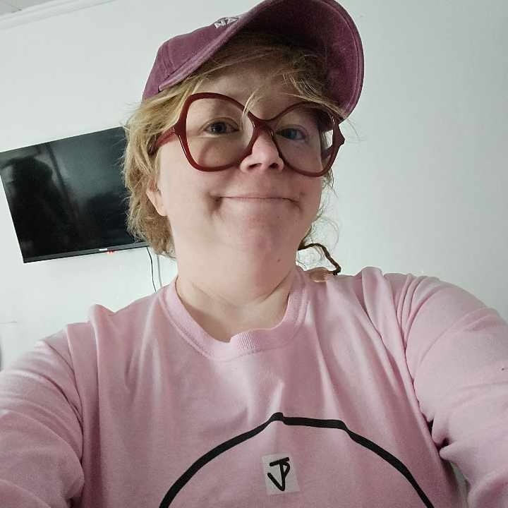 Author Rebekah Palmer uses she/they pronouns and is a chubby white woman/non binary human. She has short, blonde hair with a smallish braid on her left side. She wears butterfly-shaped red-rimmed glasses over her blue eyes. She is wearing her "You Are On Native Land" baseball cap from Urban Native Era . She is wearing her long-sleeved pink baseball shirt that has her youngest brother