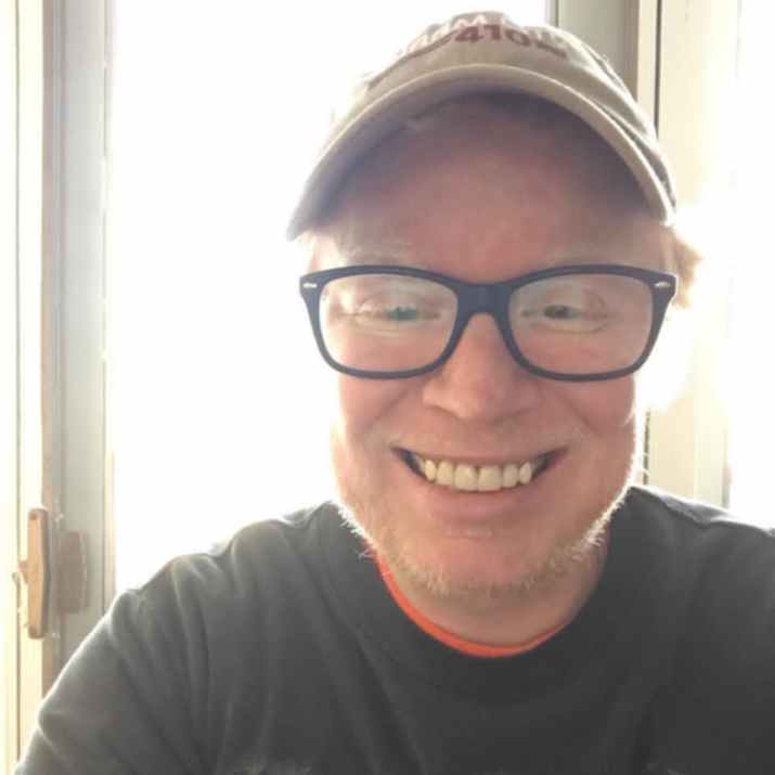 Steve is smiling into the camera. He has blonde hair and blue eyes and is wearing glasses with thick, dark frames. His shirt is green. He's standing in front of an open door with light glowing around him. This photo was taken at his home.